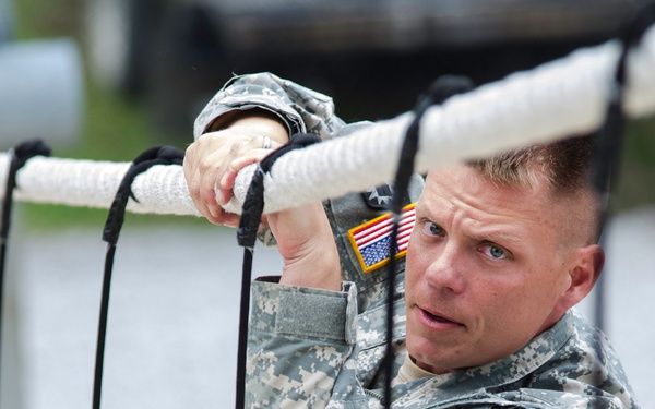 A U.S. Army soldier in Army Combat Uniform (ACU) digital camouflage pattern is navigating what appears to be a rope obstacle course or cargo net. He is gripping a white horizontal rope with both hands while black vertical ropes frame the foreground. The soldier displays focused determination and wears visible unit patches including a U.S. flag patch on his left shoulder.