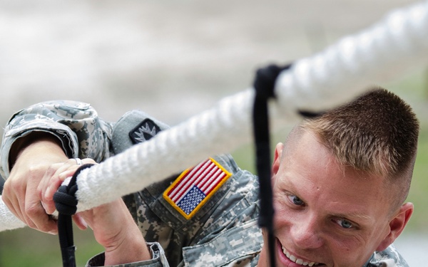 A U.S. Army soldier in Army Combat Uniform (ACU) is climbing or gripping a thick white rope while smiling. The soldier wears a reverse American flag patch on the right shoulder and an additional unit patch on the left sleeve. The image appears to be taken during a physical training or obstacle course exercise.