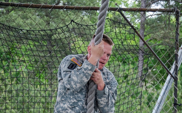 A U.S. Army soldier in ACU (Army Combat Uniform) digital camouflage is performing a rope climb exercise on a military obstacle course. The soldier is gripping a thick knotted rope while standing on a wooden platform, appearing to prepare for or demonstrate the climb. Blue safety mats are visible below, and a cargo net structure forms the backdrop.