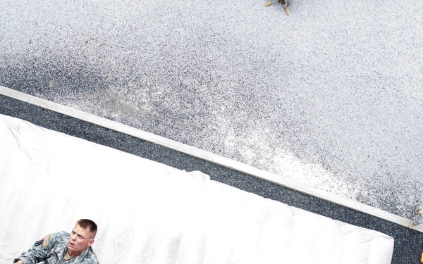 An aerial/elevated view looking down at a military soldier in camouflage uniform standing near or on a large white-tarp-covered vehicle or cargo container. Thick rope rigging is visible in the foreground, suggesting the photo was taken from an elevated structure or vehicle. Additional military personnel are visible in the background on what appears to be a wet or snow-dusted surface.