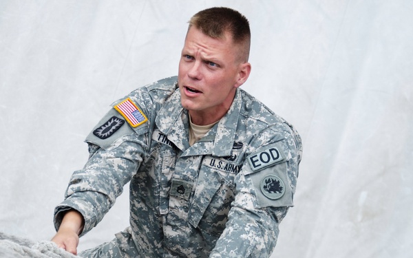 A U.S. Army soldier wearing ACU (Army Combat Uniform) with EOD (Explosive Ordnance Disposal) insignia is navigating what appears to be an obstacle course or training exercise. The soldier is climbing or crawling over a large white surface, possibly a tarp, net, or inflatable obstacle. His expression suggests physical exertion and concentration.