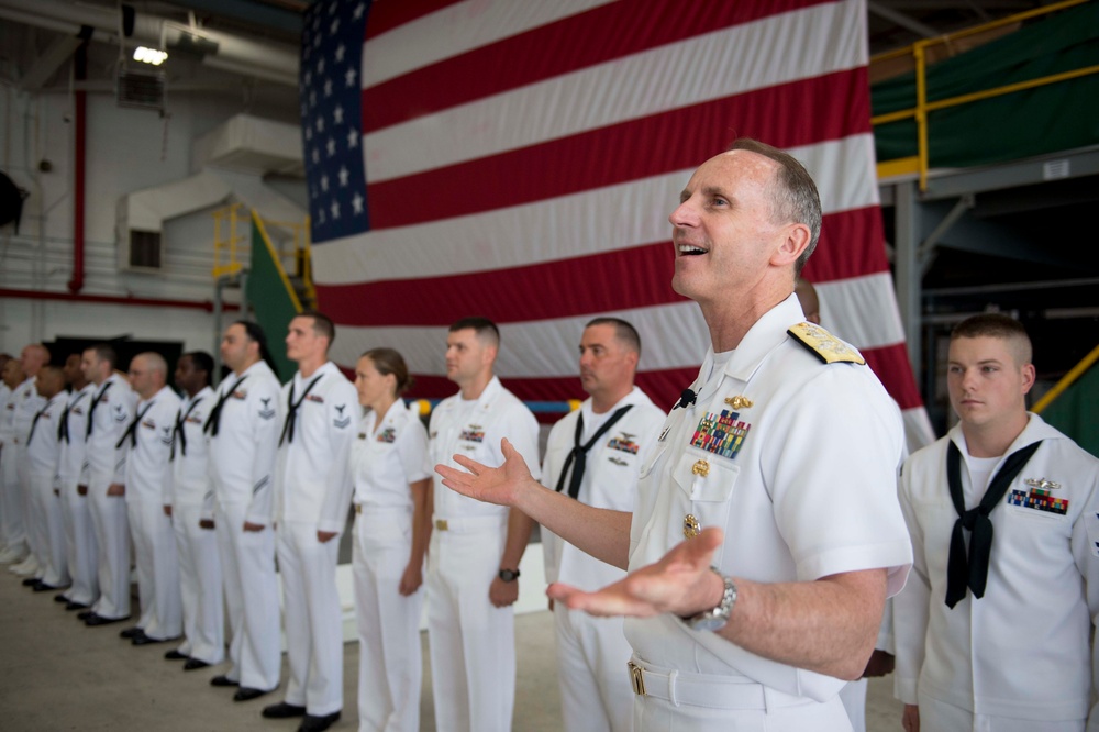 A senior U.S. Navy admiral in white dress uniform addresses a formation of sailors standing at attention in what appears to be a hangar bay or large indoor military facility. A large American flag is prominently displayed on the wall behind the formation. The admiral appears to be speaking or giving remarks, with his hands gesturing expressively.