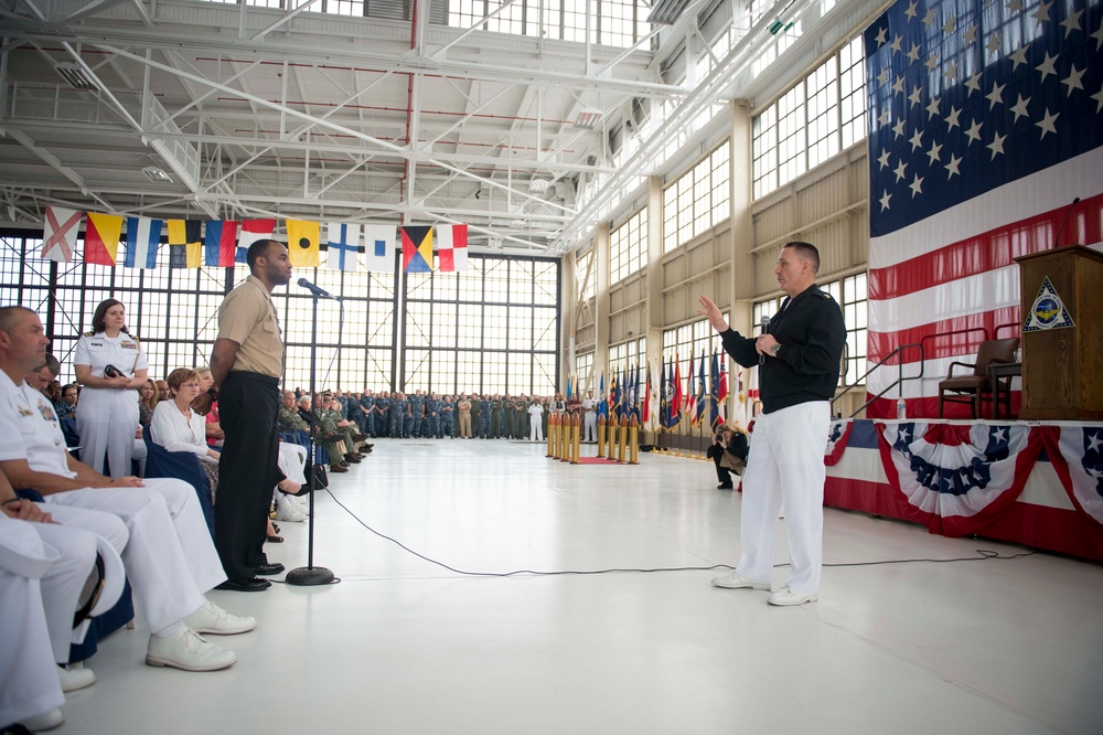 A formal U.S. military ceremony is taking place inside a large hangar facility. A senior officer in a dark top and white trousers is speaking into a microphone while addressing a service member standing at attention near a podium microphone. A large American flag serves as a backdrop on a decorated stage, with rows of military personnel and civilians seated and standing as an audience.