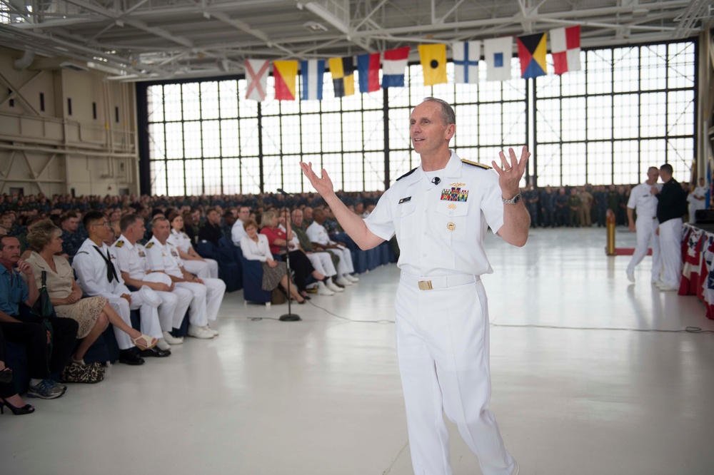 A senior naval officer in white dress uniform is addressing a large audience inside a military hangar facility. The officer is gesturing with both hands raised while speaking to a crowd of uniformed military personnel and civilians seated in rows. International maritime signal flags are displayed across the hangar's large windows in the background.