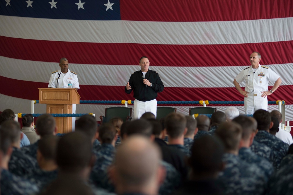 A military all-hands or command briefing is taking place in a large indoor venue, likely a hangar bay, with a large American flag displayed as a backdrop. Three uniformed military officers are on stage — two in white Navy dress uniforms and one in a dark Navy working uniform speaking into a microphone. A large audience of sailors in blue digital camouflage uniforms is seated facing the stage.