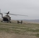 A military medical evacuation (MEDEVAC) scene showing soldiers carrying a litter/stretcher toward a landed helicopter in an arid, mountainous terrain. The helicopter appears to be a Mi-17 or similar Soviet-era transport helicopter in military camouflage livery. The overcast conditions and sparse landscape suggest a Central Asian or Middle Eastern operational environment.