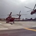 Two military helicopters are visible on what appears to be a military airfield or helipad. One helicopter is on the ground with rotors spinning, while a second identical helicopter is airborne nearby, likely landing or departing. Both aircraft appear to be painted in a desert/sand camouflage color scheme consistent with Mi-17 or similar Soviet-era transport helicopters.