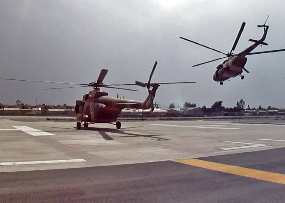 Two military helicopters are visible on what appears to be a military airfield or helipad. One helicopter is on the ground with rotors spinning, while a second identical helicopter is airborne nearby, likely landing or departing. Both aircraft appear to be painted in a desert/sand camouflage color scheme consistent with Mi-17 or similar Soviet-era transport helicopters.