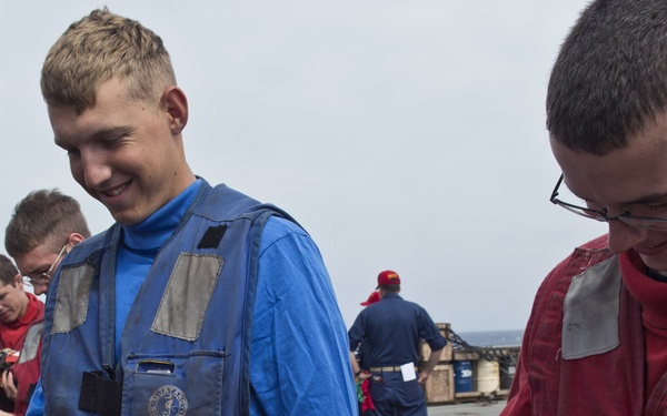 Two young personnel in blue and red flight deck jerseys/vests are examining aviation cranial helmets on what appears to be a naval vessel's flight deck. The individual on the right has a name tag reading 'STEARNS' visible on his red vest. Several other personnel in similar colored vests are visible in the background.
