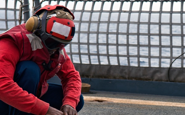 A naval crew member wearing a red jersey and protective helmet with ear defenders is crouched on what appears to be a ship's flight deck, handling a tan/beige tie-down strap marked with the number '0412'. The individual is working near safety netting at the deck's edge with open ocean visible in the background. The red jersey indicates a specific role designation typical of U.S. Navy carrier deck operations.