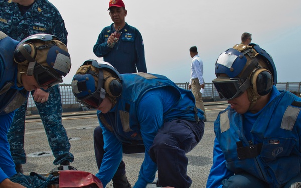 U.S. Navy personnel in blue flight deck gear and helmets conduct a medical casualty evacuation drill on what appears to be a ship's flight deck. Three crew members in blue coveralls and cranial helmets are positioning a training mannequin dressed in Navy digital camouflage uniform onto a wire Stokes litter stretcher. Observers in Navy working uniforms, including one wearing a red cap, watch the exercise from the background.