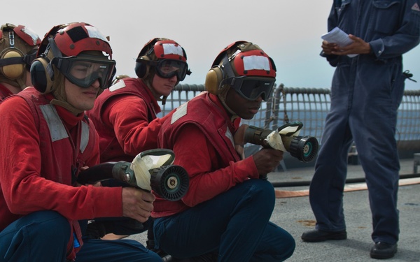 Several naval personnel dressed in red jerseys, protective helmets with goggles, and hearing protection are crouching on what appears to be a ship's flight deck. They are holding firefighting nozzles in a ready position, suggesting a damage control or firefighting drill. A supervisor in blue coveralls stands nearby holding papers, observing the exercise.