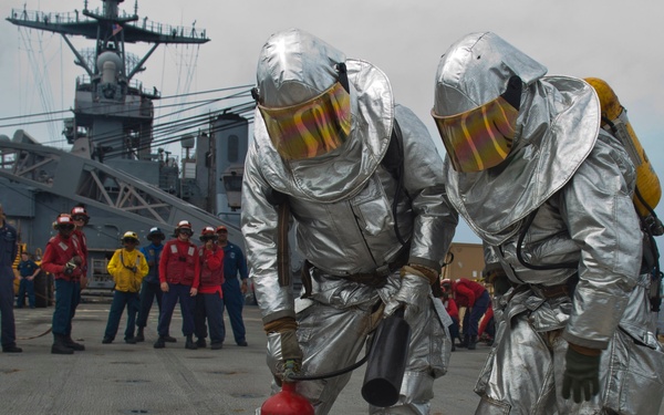 Two personnel in silver aluminized proximity/fire proximity suits with yellow visors and yellow boots are handling a red fire extinguisher on the deck of a naval vessel. A group of observers in various colored uniforms (red, yellow, blue) watch from the background, suggesting this is a firefighting drill or training exercise. A large naval warship superstructure is visible in the background.