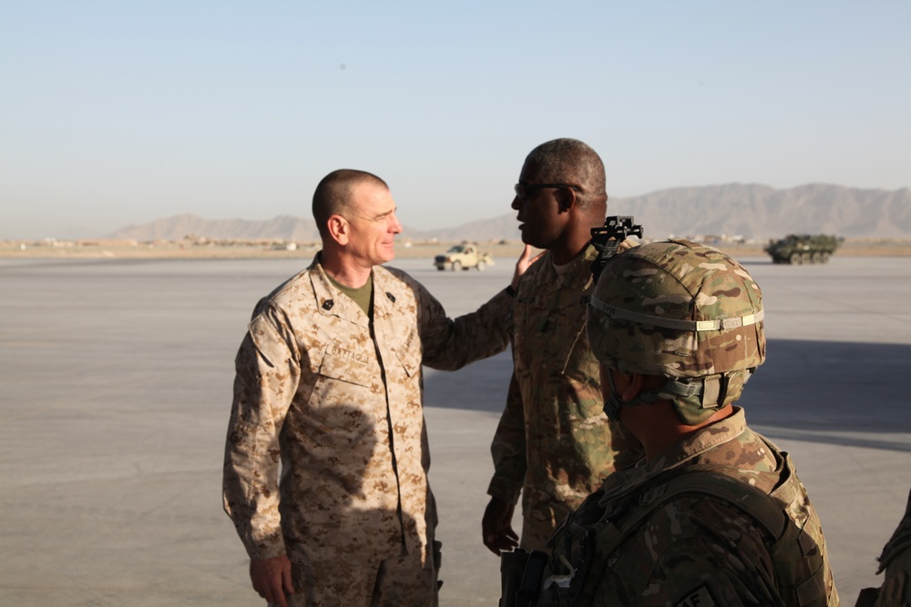 Military personnel in different camouflage uniforms are interacting on what appears to be a tarmac or airfield in an arid, mountainous environment. One individual wearing USMC MARPAT desert camouflage is engaged in conversation with soldiers wearing OCP (Operational Camouflage Pattern) uniforms. Military vehicles are visible in the background.