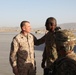 Military personnel in different camouflage uniforms are interacting on what appears to be a tarmac or airfield in an arid, mountainous environment. One individual wearing USMC MARPAT desert camouflage is engaged in conversation with soldiers wearing OCP (Operational Camouflage Pattern) uniforms. Military vehicles are visible in the background.