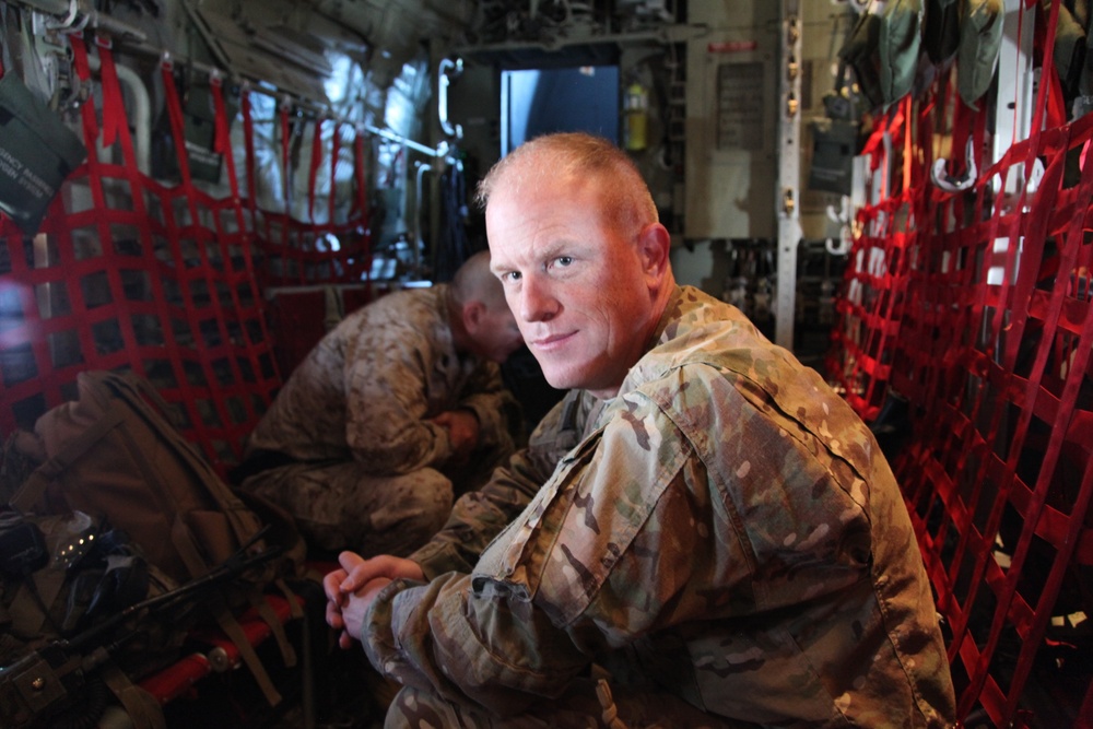 A military service member in MultiCam/OCP camouflage uniform is seated inside what appears to be a military transport aircraft, likely a C-130 or similar cargo plane, identifiable by the characteristic red cargo netting on the fuselage walls. He is looking directly at the camera with a relaxed posture, hands clasped. A second service member in a different camouflage pattern is visible in the background, head down.