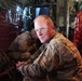 A military service member in MultiCam/OCP camouflage uniform is seated inside what appears to be a military transport aircraft, likely a C-130 or similar cargo plane, identifiable by the characteristic red cargo netting on the fuselage walls. He is looking directly at the camera with a relaxed posture, hands clasped. A second service member in a different camouflage pattern is visible in the background, head down.