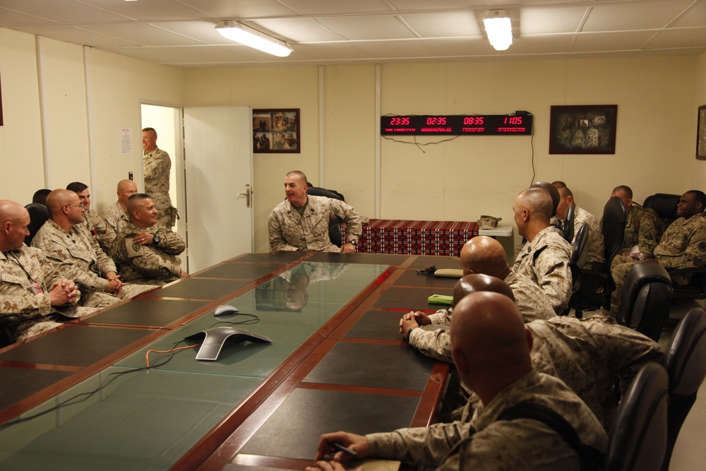 Military personnel in camouflage uniforms are gathered around a large conference table in what appears to be a forward operating base briefing room. A multi-timezone digital clock display on the wall shows four different times (23:35, 02:35, 08:35, 11:05), indicating a multinational or multi-theater operational context. The group appears to be engaged in an informal discussion or briefing.