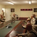 Military personnel in camouflage uniforms are gathered around a large conference table in what appears to be a forward operating base briefing room. A multi-timezone digital clock display on the wall shows four different times (23:35, 02:35, 08:35, 11:05), indicating a multinational or multi-theater operational context. The group appears to be engaged in an informal discussion or briefing.