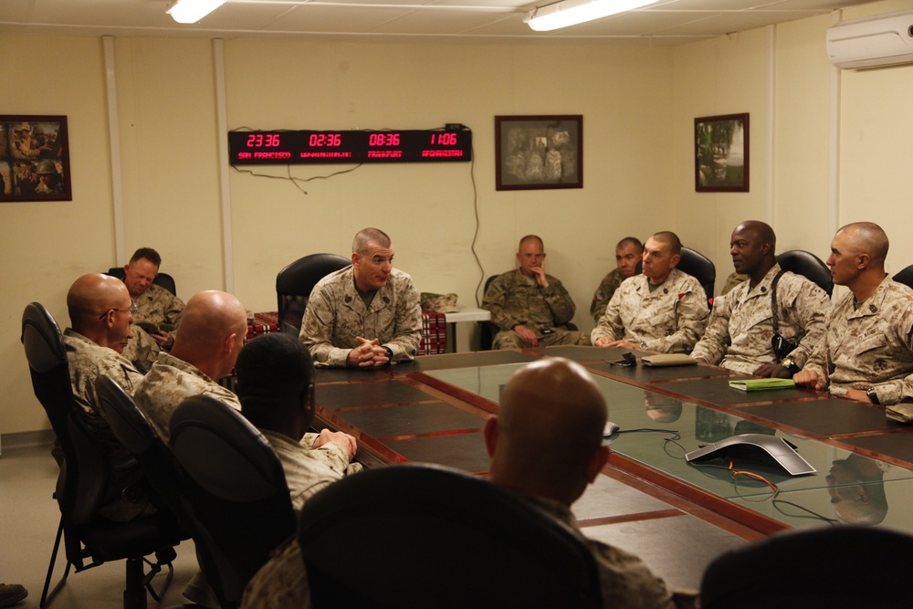 A military briefing or meeting is taking place in a conference room, with approximately 10-12 personnel in desert camouflage uniforms seated around a large table. A multi-timezone digital clock display on the wall shows times for San Francisco, Washington D.C., Frankfurt, and Afghanistan, indicating this is likely a forward operating base or command facility. One individual appears to be leading the discussion from the far side of the table.