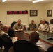 A military briefing or meeting is taking place in a conference room, with approximately 10-12 personnel in desert camouflage uniforms seated around a large table. A multi-timezone digital clock display on the wall shows times for San Francisco, Washington D.C., Frankfurt, and Afghanistan, indicating this is likely a forward operating base or command facility. One individual appears to be leading the discussion from the far side of the table.