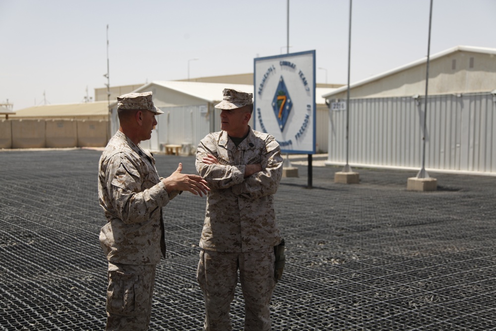 Two U.S. Marine Corps personnel in desert MARPAT camouflage uniforms are engaged in conversation at what appears to be a forward operating base in an arid environment. A sign in the background displays a Marine Corps emblem with the number '7', consistent with Marine Combat Team 7 or a similar unit designation. The setting includes temporary structures, T-barriers, and a gravel/matting surface typical of deployed military installations.