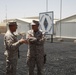 Two U.S. Marine Corps personnel in desert MARPAT camouflage uniforms are engaged in conversation at what appears to be a forward operating base in an arid environment. A sign in the background displays a Marine Corps emblem with the number '7', consistent with Marine Combat Team 7 or a similar unit designation. The setting includes temporary structures, T-barriers, and a gravel/matting surface typical of deployed military installations.