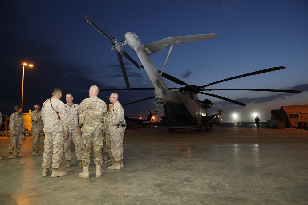 A group of military personnel in camouflage uniforms are gathered on a flight line or tarmac at dusk or dawn, engaged in conversation near a large military helicopter. The helicopter appears to be a heavy-lift rotary-wing aircraft, likely a CH-53 Super Stallion or similar platform, with its rotor blades visible overhead. The setting appears to be a forward operating base or military airfield.