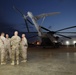 A group of military personnel in camouflage uniforms are gathered on a flight line or tarmac at dusk or dawn, engaged in conversation near a large military helicopter. The helicopter appears to be a heavy-lift rotary-wing aircraft, likely a CH-53 Super Stallion or similar platform, with its rotor blades visible overhead. The setting appears to be a forward operating base or military airfield.
