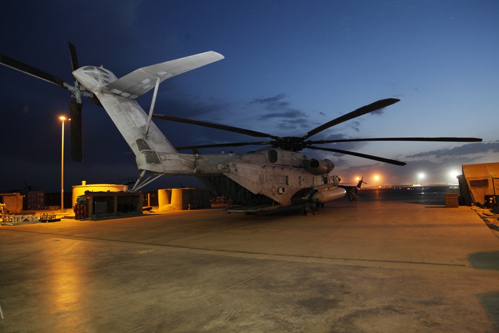 A large U.S. Marines heavy-lift helicopter, identifiable as a Sikorsky CH-53E Super Stallion, is parked on a concrete flight line apron at dusk or dawn. The aircraft's tail rotor assembly is folded upward, indicating a space-saving configuration typical of shipboard or forward operating base storage. The scene is illuminated by artificial lighting against a deep blue twilight sky.