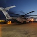 A large U.S. Marines heavy-lift helicopter, identifiable as a Sikorsky CH-53E Super Stallion, is parked on a concrete flight line apron at dusk or dawn. The aircraft's tail rotor assembly is folded upward, indicating a space-saving configuration typical of shipboard or forward operating base storage. The scene is illuminated by artificial lighting against a deep blue twilight sky.