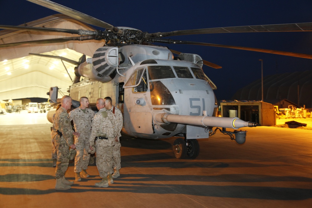 A group of military personnel in desert camouflage uniforms are gathered on a flight line at night, conversing in front of a large military helicopter marked with the number '51'. The aircraft appears to be a Sikorsky CH-53 Sea Stallion or similar heavy-lift helicopter, consistent with U.S. Marine Corps markings. The scene is set on a military airfield with an illuminated hangar structure visible in the background.