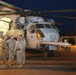 A group of military personnel in desert camouflage uniforms are gathered on a flight line at night, conversing in front of a large military helicopter marked with the number '51'. The aircraft appears to be a Sikorsky CH-53 Sea Stallion or similar heavy-lift helicopter, consistent with U.S. Marine Corps markings. The scene is set on a military airfield with an illuminated hangar structure visible in the background.