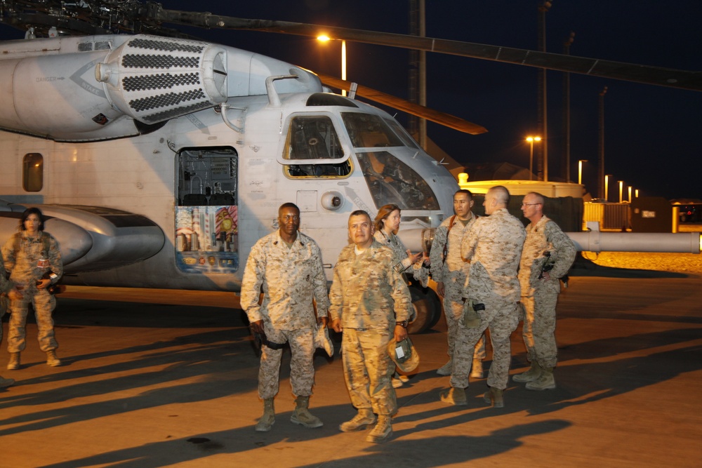 A group of military personnel in desert camouflage uniforms are gathered on a tarmac at night in front of a large military helicopter, likely a CH-53 or similar heavy-lift helicopter. The scene appears to be at a forward operating base or military airfield, illuminated by artificial lighting. Several service members appear to have just disembarked or are preparing to board the aircraft.