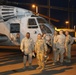 A group of military personnel in desert camouflage uniforms are gathered on a tarmac at night in front of a large military helicopter, likely a CH-53 or similar heavy-lift helicopter. The scene appears to be at a forward operating base or military airfield, illuminated by artificial lighting. Several service members appear to have just disembarked or are preparing to board the aircraft.