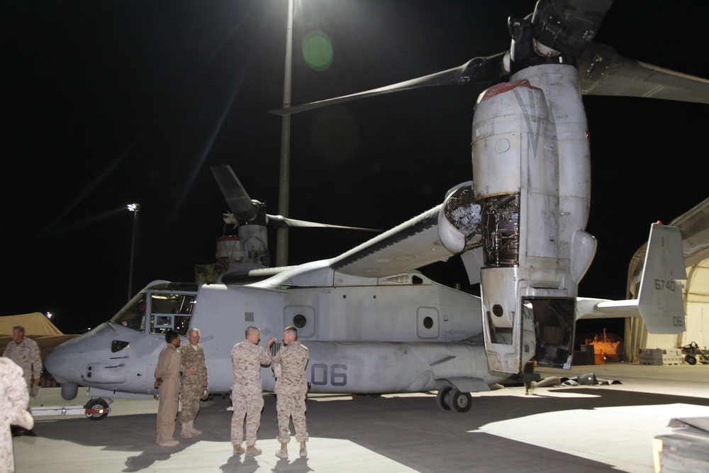 A nighttime military scene showing a V-22 Osprey tiltrotor aircraft parked on a flight line or tarmac. Several military personnel in various uniforms are gathered near the nose of the aircraft, appearing to be in conversation or conducting a briefing. The aircraft's starboard engine nacelle is open, suggesting maintenance or an inspection is underway.