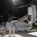 A nighttime military scene showing a V-22 Osprey tiltrotor aircraft parked on a flight line or tarmac. Several military personnel in various uniforms are gathered near the nose of the aircraft, appearing to be in conversation or conducting a briefing. The aircraft's starboard engine nacelle is open, suggesting maintenance or an inspection is underway.