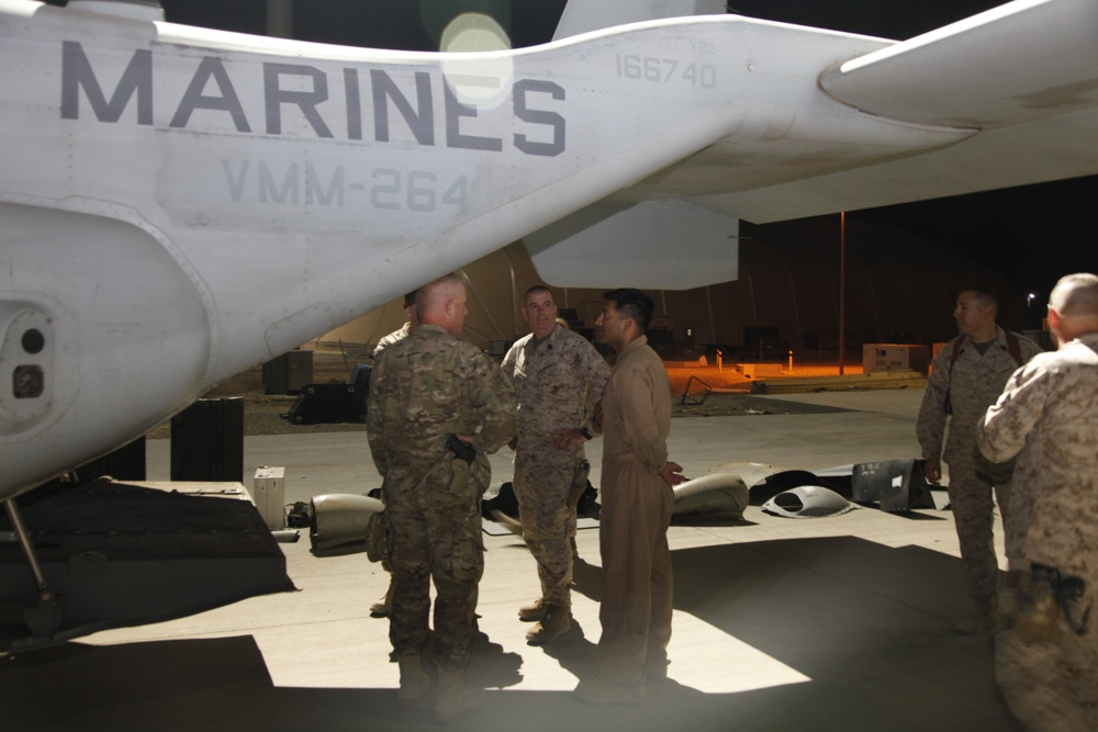 Military personnel are gathered on a flight line at night near a Marine Corps aircraft bearing the markings 'MARINES', 'VMM-264', and bureau number '166740'. Several individuals in various military uniforms appear to be engaged in conversation or a briefing near the aircraft. The setting appears to be a forward operating base or air station flight line.