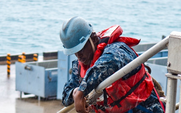 A sailor in U.S. Navy blue digital camouflage (NWU Type I) uniform is working on the deck of a naval vessel in rainy conditions. The individual is wearing full safety gear including a blue hard hat, red inflatable life vest, and a blue safety harness, and appears to be manipulating a fitting or clamp on a metal railing. The wet deck and open water visible in the background indicate active shipboard operations at sea or near a pier.