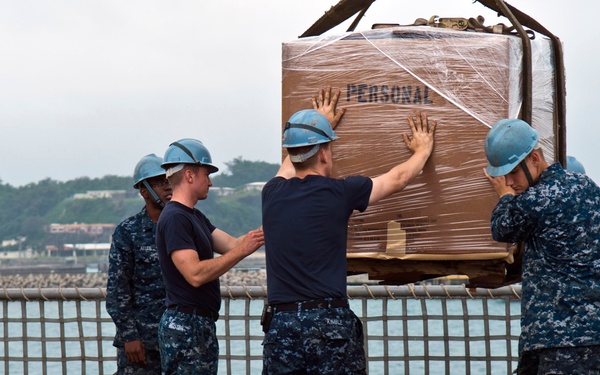 A group of U.S. Navy sailors in digital camouflage uniforms and blue hard hats are handling a large palletized cargo load labeled 'PERSONAL' on what appears to be a ship's deck. The pallet is wrapped in plastic and is being guided or steadied by multiple personnel, suggesting it is being lowered or positioned via crane. The scene appears to take place in an overseas port, with water and a coastal installation visible in the background.