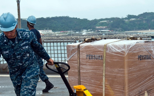 A U.S. Navy sailor in blue digital camouflage (NWU) and a blue hard hat is pulling a yellow pallet jack loaded with a large shrink-wrapped pallet marked 'PERSONAL' on a wet deck surface. The setting appears to be a naval pier or ship deck at what looks like a port facility, with hills and harbor infrastructure visible in the background. A second sailor in similar uniform is visible in the background.