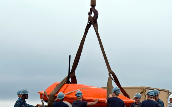 A group of approximately eight to nine military personnel in U.S. Navy digital camouflage uniforms are working on a ship's deck to handle a bright orange aerial target drone being lifted by a crane with rigging straps. The personnel are wearing hard hats and coordinating the positioning of the drone onto what appears to be a launch rail or cradle. The scene takes place on a wet ship deck under overcast skies with the ocean visible in the background.