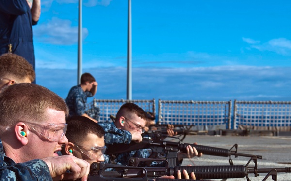 A group of military personnel in U.S. Navy digital camouflage (NWU Type I) uniforms are conducting a rifle qualification exercise on what appears to be the deck of a naval vessel. The personnel are in the prone position aiming M16/M4-style rifles downrange. All visible shooters are wearing safety glasses and ear protection.