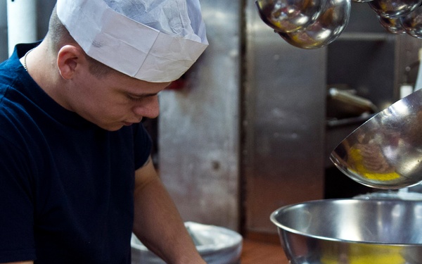 A male kitchen worker wearing a white paper chef's hat and navy blue shirt is preparing raw sausages on a yellow cutting board placed on a metal baking tray. The setting is a commercial kitchen with stainless steel equipment visible in the background. The worker appears to be arranging or handling the sausages in preparation for cooking.