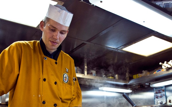 A cook or culinary specialist wearing a mustard-yellow chef's jacket with an embroidered patch and a white paper toque is working in a professional kitchen. He is spreading or leveling white frosting/cream on a sheet cake placed on a baking tray. The kitchen environment appears to be a military or institutional galley, indicated by the stainless steel surroundings and the uniform patch.