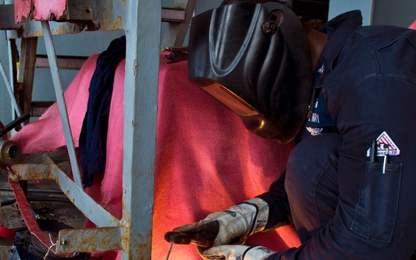 A welder in full protective gear is performing welding work at the base of a metal structural support, crouching close to the floor. The worker is surrounded by fire-resistant welding blankets (red/pink) to contain sparks, with visible welding sparks and bright light at the weld point. Safety equipment including a welding helmet, heavy gloves, and flame-resistant coveralls are clearly visible.