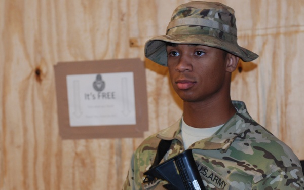 A young U.S. Army soldier in Operational Camouflage Pattern (OCP) uniform stands at what appears to be an indoor military facility or forward operating base. He is wearing a boonie hat and carrying a rifle, appearing to be on guard duty or security detail. Small American flags and a sign reading 'It's FREE' are visible in the background near a table.