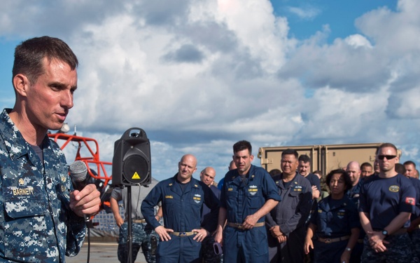 A U.S. Navy officer identified by name tape as 'BARNETT' addresses a gathered group of military personnel while holding a microphone and papers. The audience consists of approximately 20-30 service members in various uniforms, standing on what appears to be a ship's deck or tarmac. The setting suggests an official military briefing or ceremony conducted outdoors.