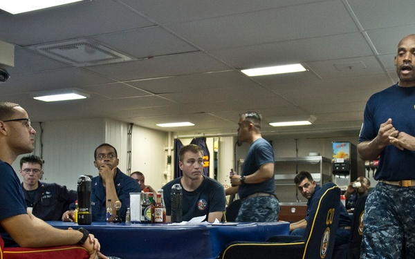A group of U.S. Navy personnel are gathered in what appears to be a ship's mess hall or briefing room. A standing sailor in Navy Working Uniform (NWU) is addressing seated personnel around a blue-clothed table. The setting suggests a formal briefing, training session, or command meeting aboard a naval vessel.