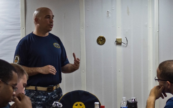 A bald man in a navy blue military uniform is standing and addressing a group of seated personnel around a blue table. The setting appears to be a briefing or meeting room aboard a naval vessel or military facility. Multiple small bottles and beverages are scattered across the table, suggesting a training or tasting exercise.